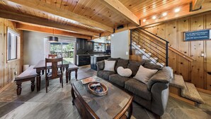 A cozy living room with a wooden ceiling, a dining area with a table and chairs, a kitchen in the background, and a staircase with a sign. - Bear-Ski Cabin: Hot Tub, Pets, BBQ (South Lake Tahoe)