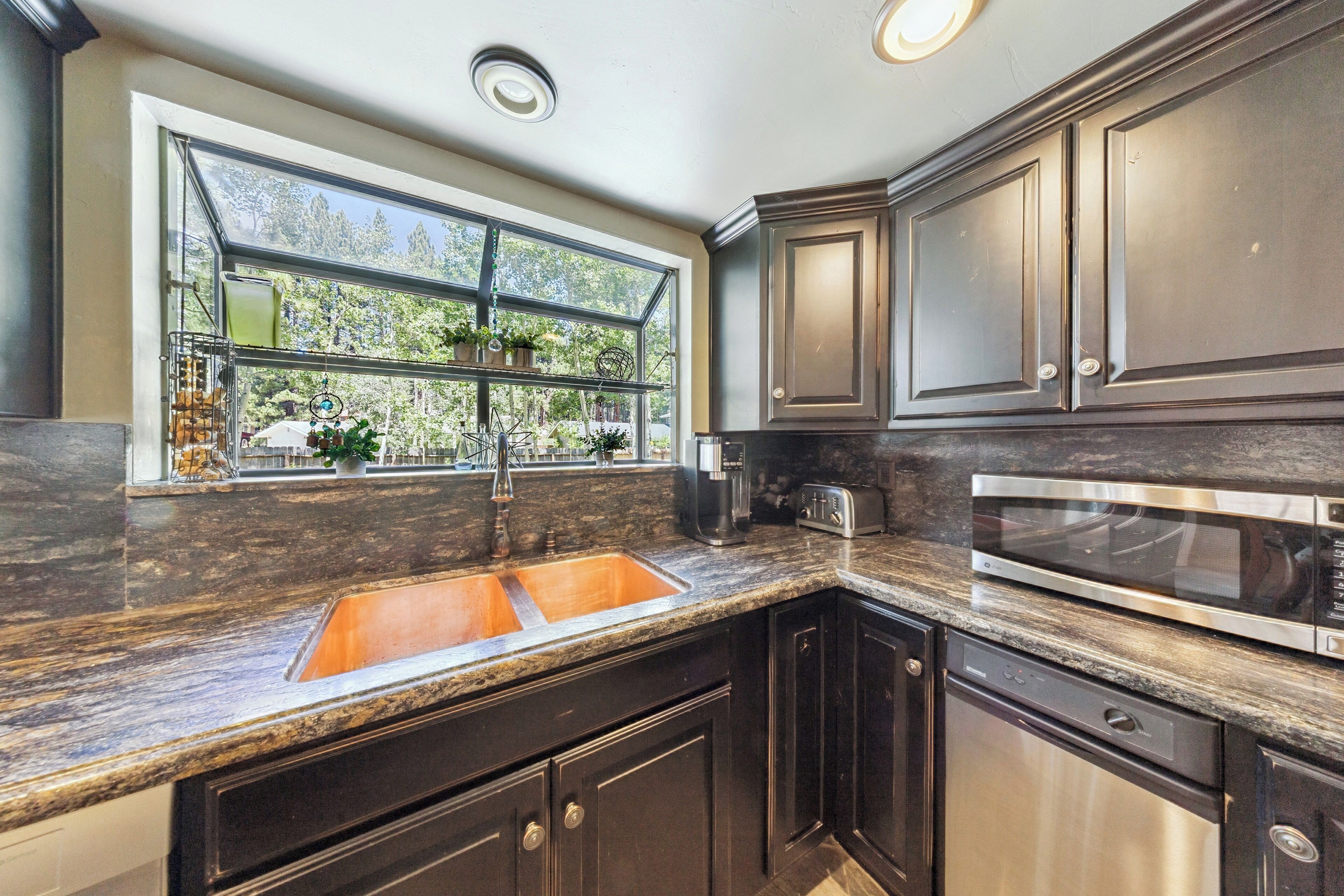 A kitchen with dark cabinetry, a stainless steel microwave, a built-in oven, a double sink, and a window with a view of trees.