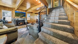 A log cabin interior with a stone fireplace, a stone staircase, and a large window overlooking a mountainous landscape. - Bear-Ski Cabin: Hot Tub, Pets, BBQ (South Lake Tahoe)