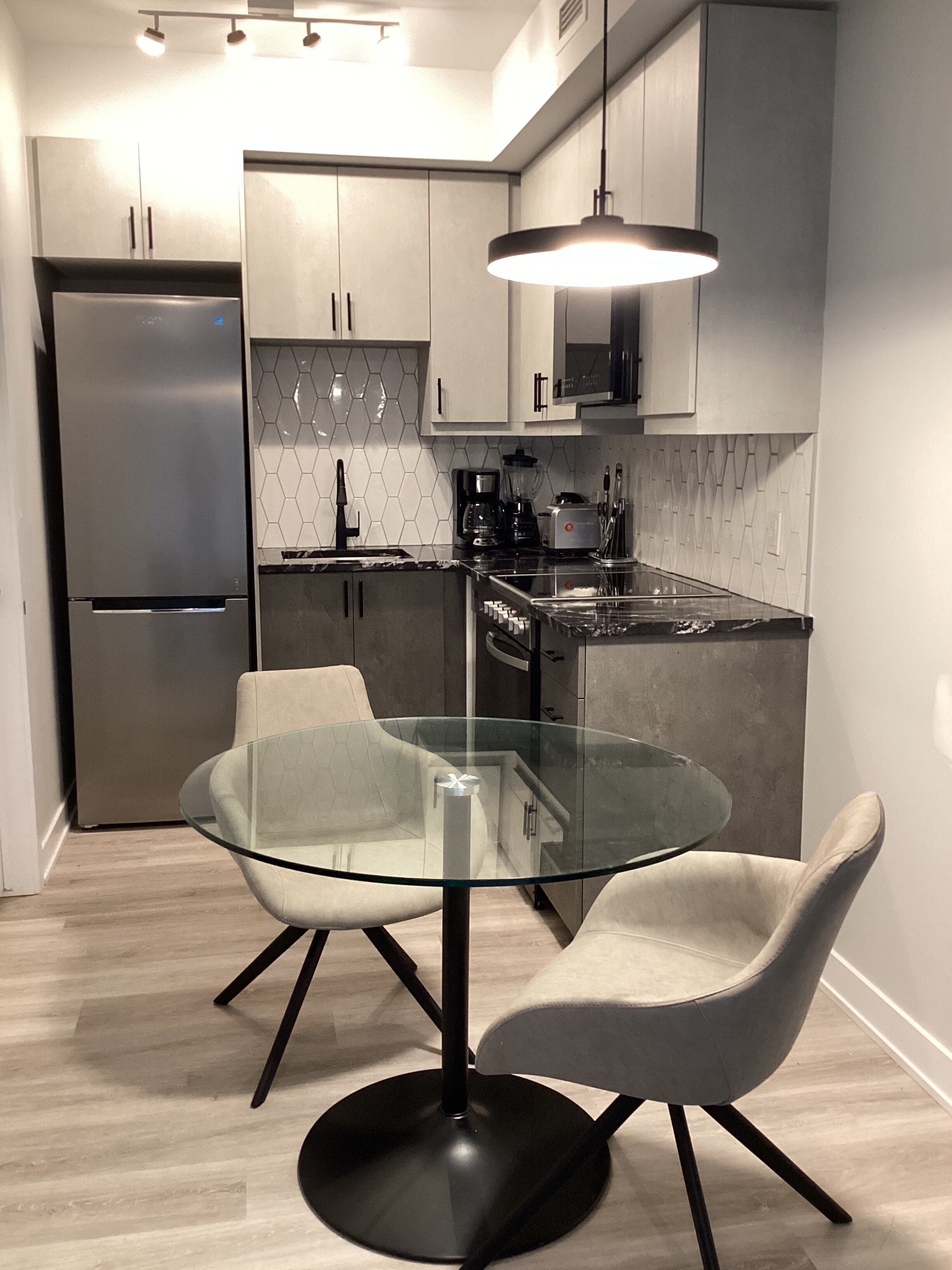 A modern kitchen with a glass-top table, two chairs, a refrigerator, a microwave, and a coffee maker.