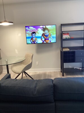 A modern living room with a flat-screen TV displaying an animated show, a glass table, a cushioned chair, and a bookshelf with books and a dictionary.
