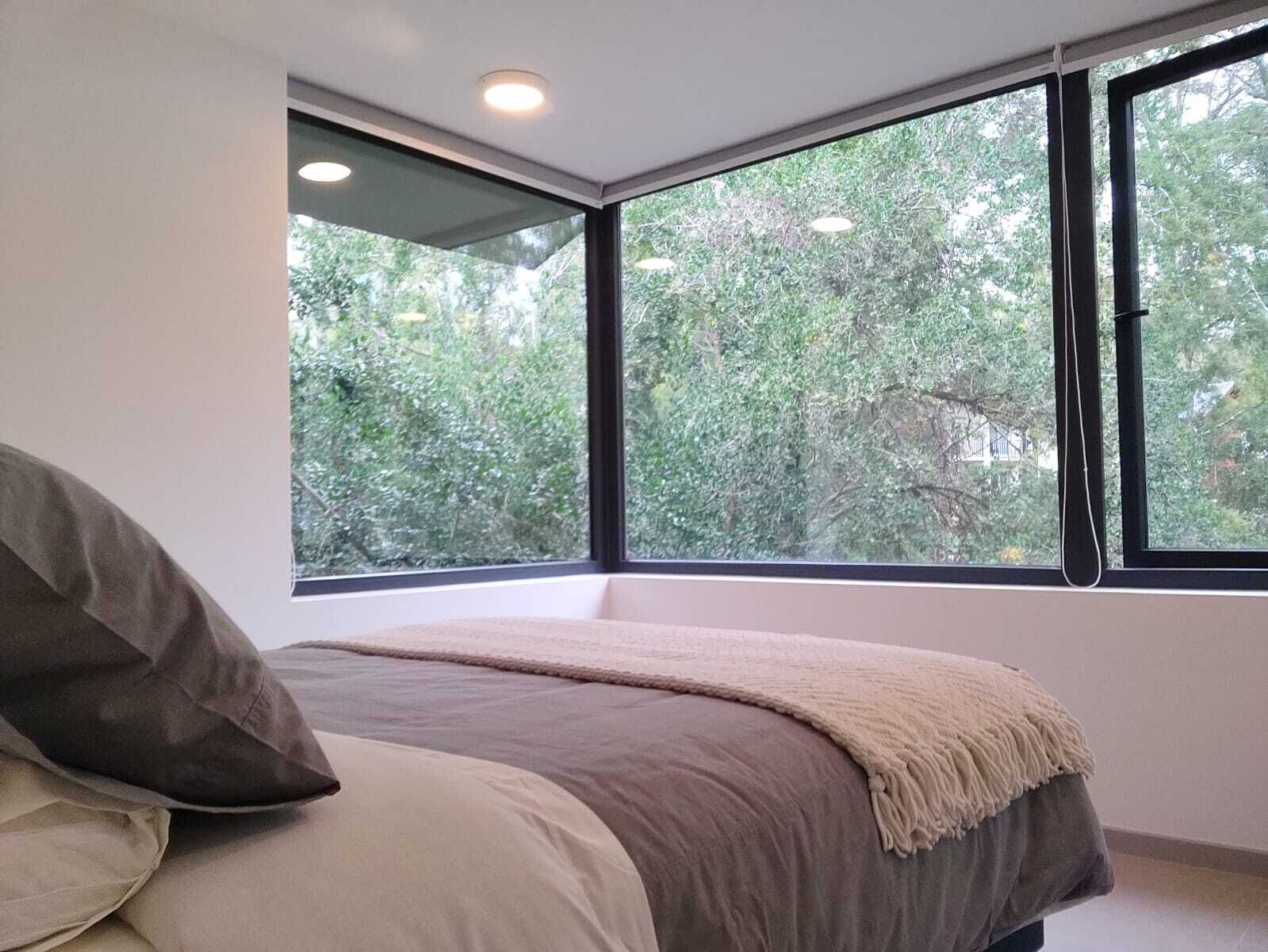 A bedroom with a large window, a bed with a beige and gray blanket, and a white wall.