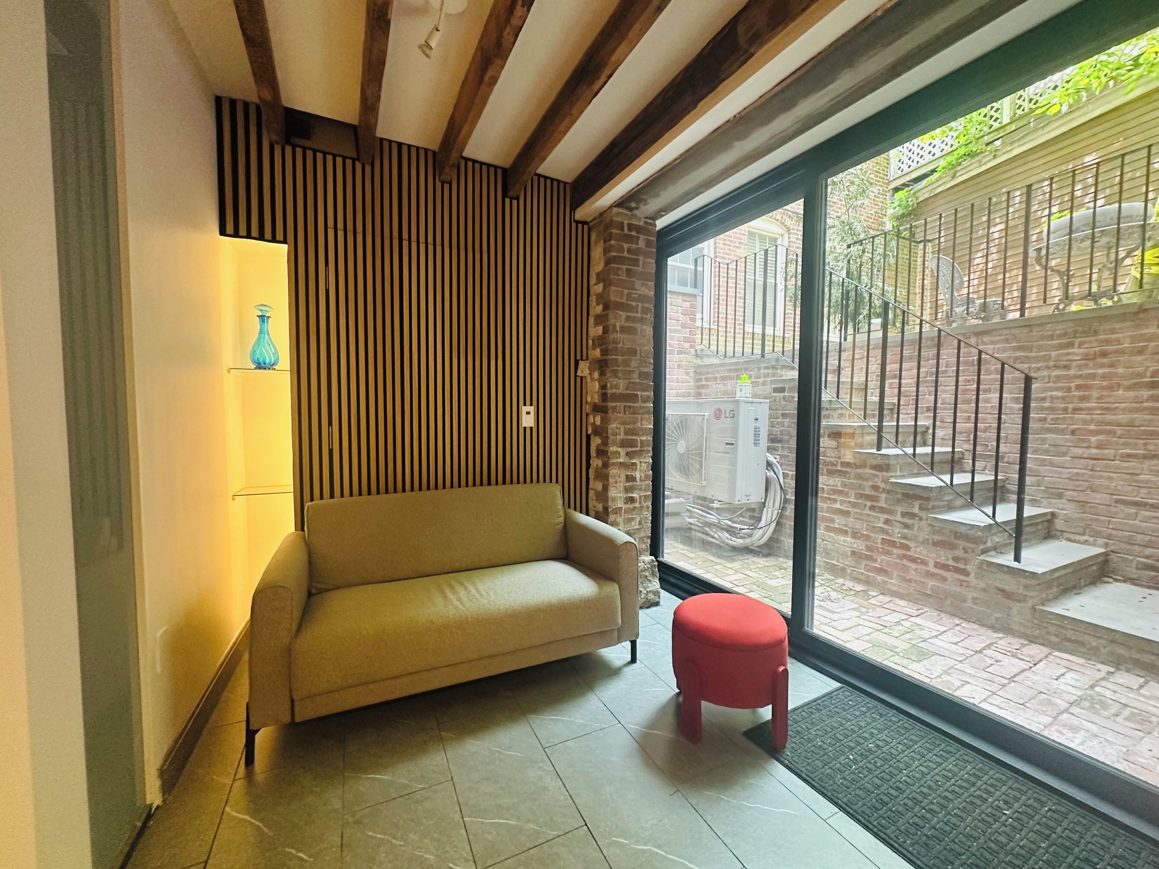 Una habitación con un sofá, un taburete pequeño, una lámpara de pared y un ventanal con vistas a una escalera de ladrillo y zona exterior.