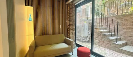 A room with a sofa, a small stool, a wall-mounted light fixture, and a large window with a view of a brick staircase and outdoor area.