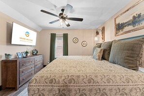 Quarto com cama, ventoinha no teto, penteadeira, televisão e um quadro na parede.