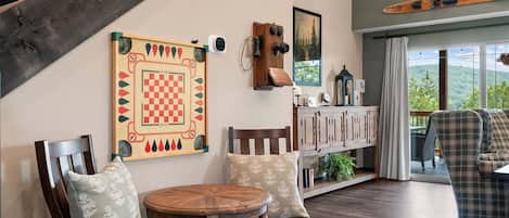 A cozy living room with a wooden table, chairs, a checkered wall hanging, and a view of the outdoors.