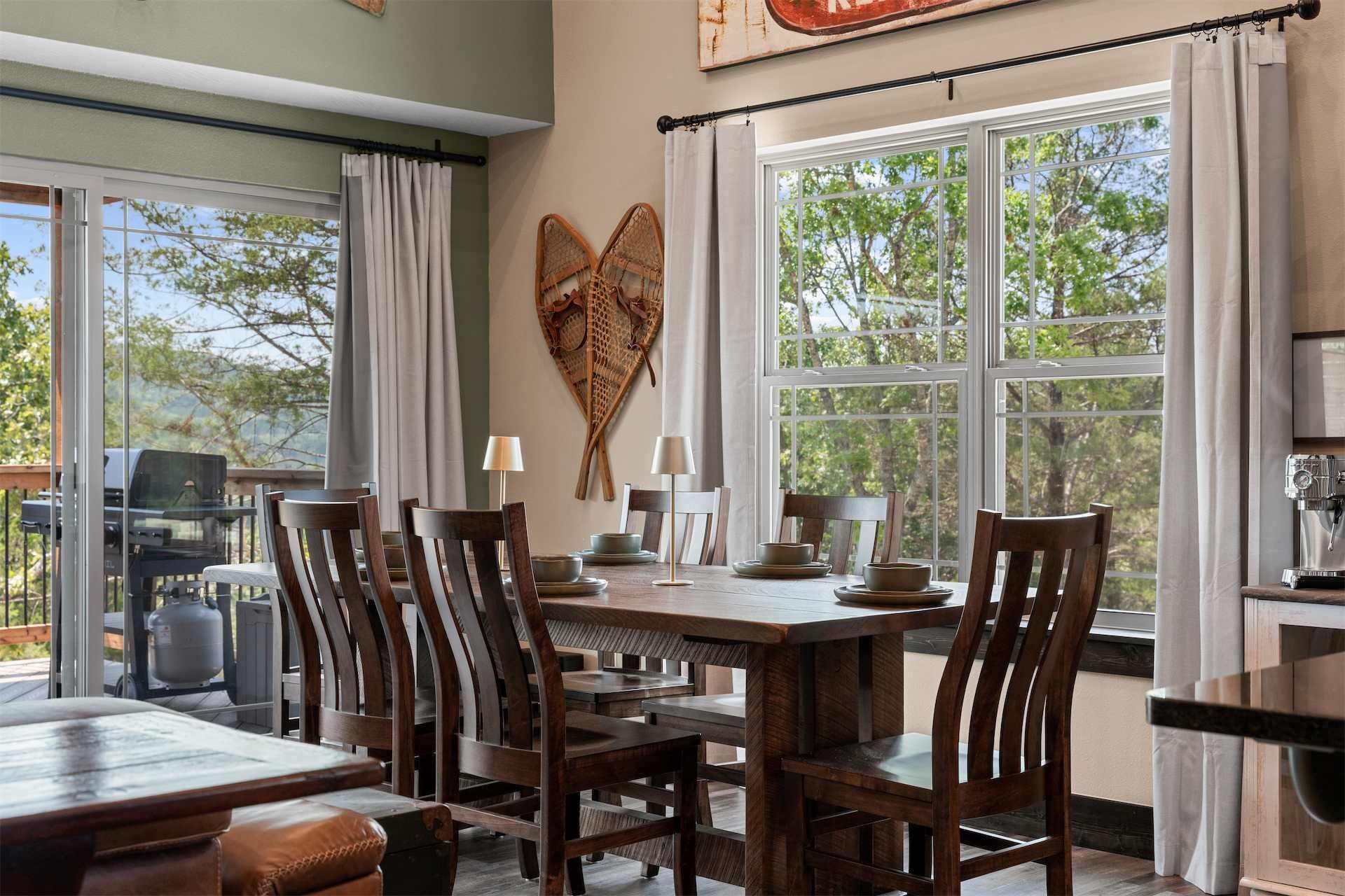 A dining area with a large table, chairs, and a view of trees through large windows.