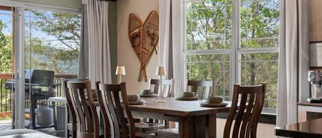 A dining area with a large table, chairs, and a view of trees through large windows.