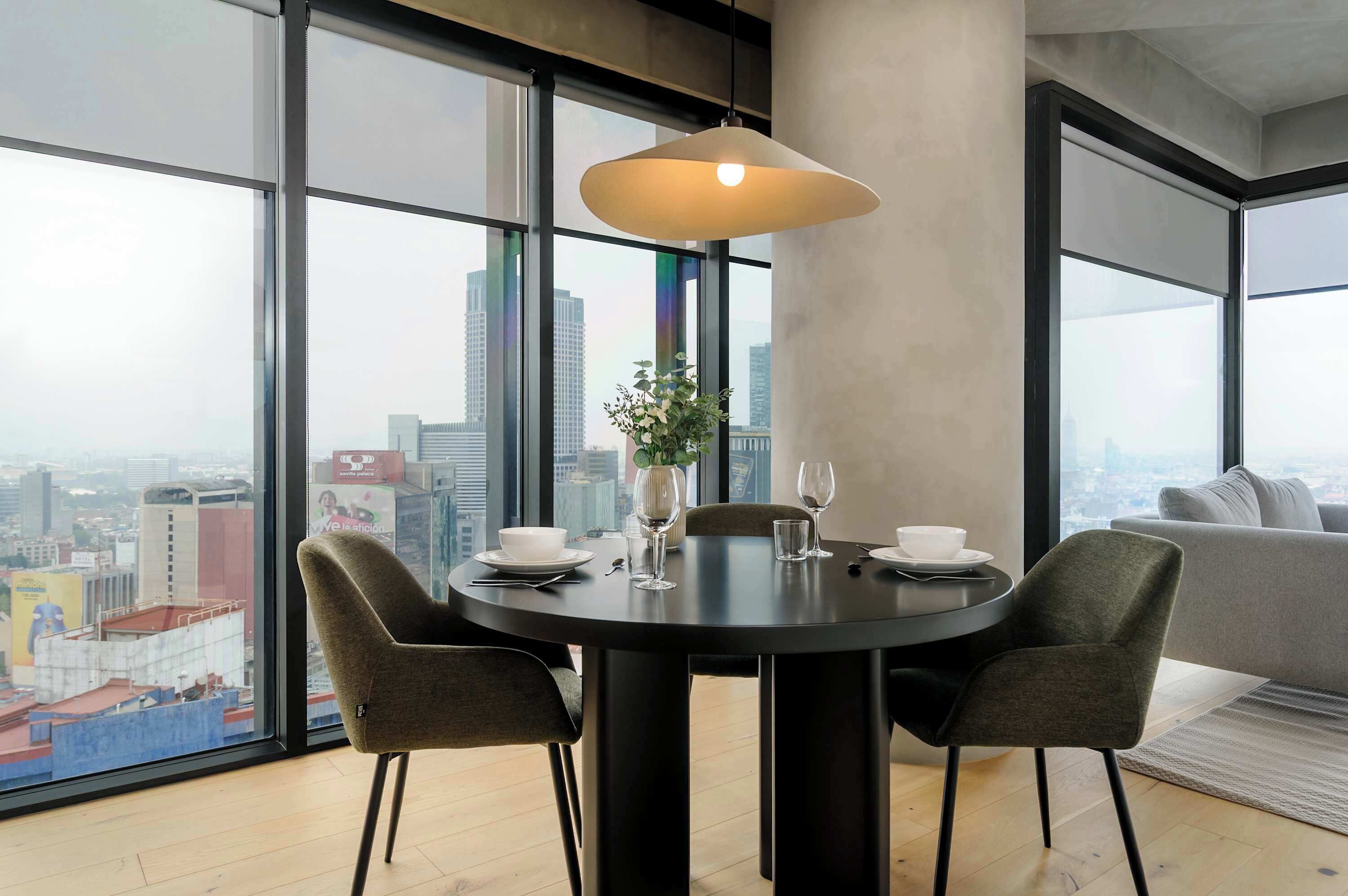 Ein stilvoller Essbereich mit einem runden Tisch, vier Stühlen, einer Vase mit Blumen und Blick auf die Stadt durch große Fenster.