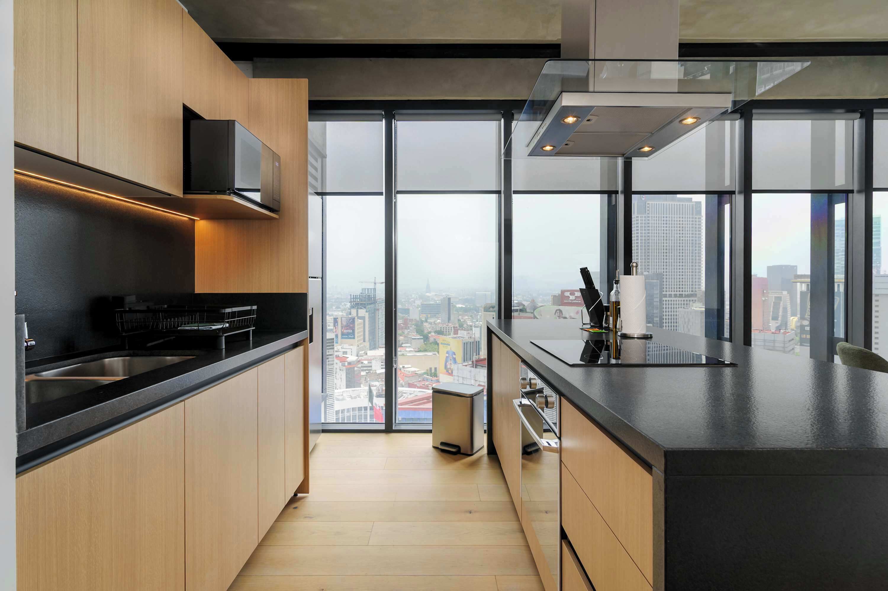 A modern kitchen with a city view, featuring a stainless steel sink, black countertop, and wooden cabinets.