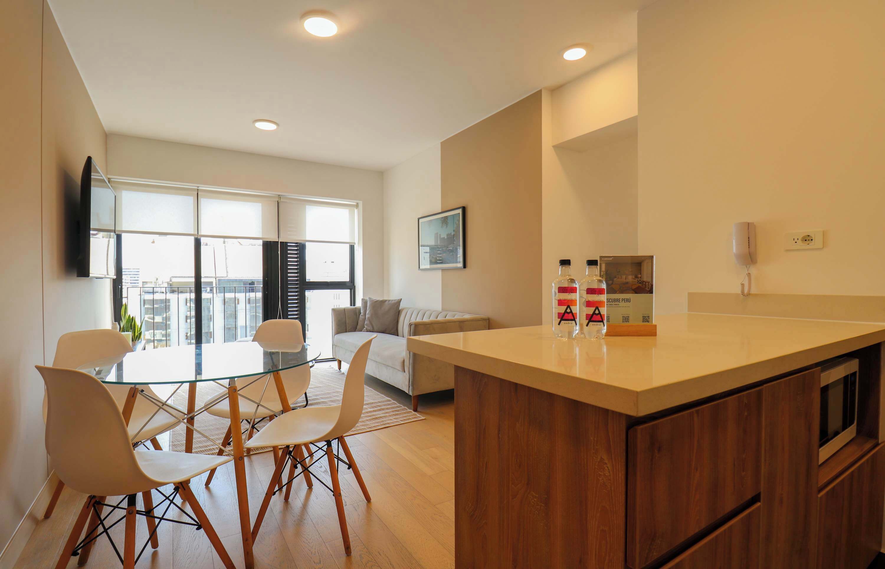A modern kitchen with a central island, dining area with chairs, a sofa, and a TV mounted on the wall.