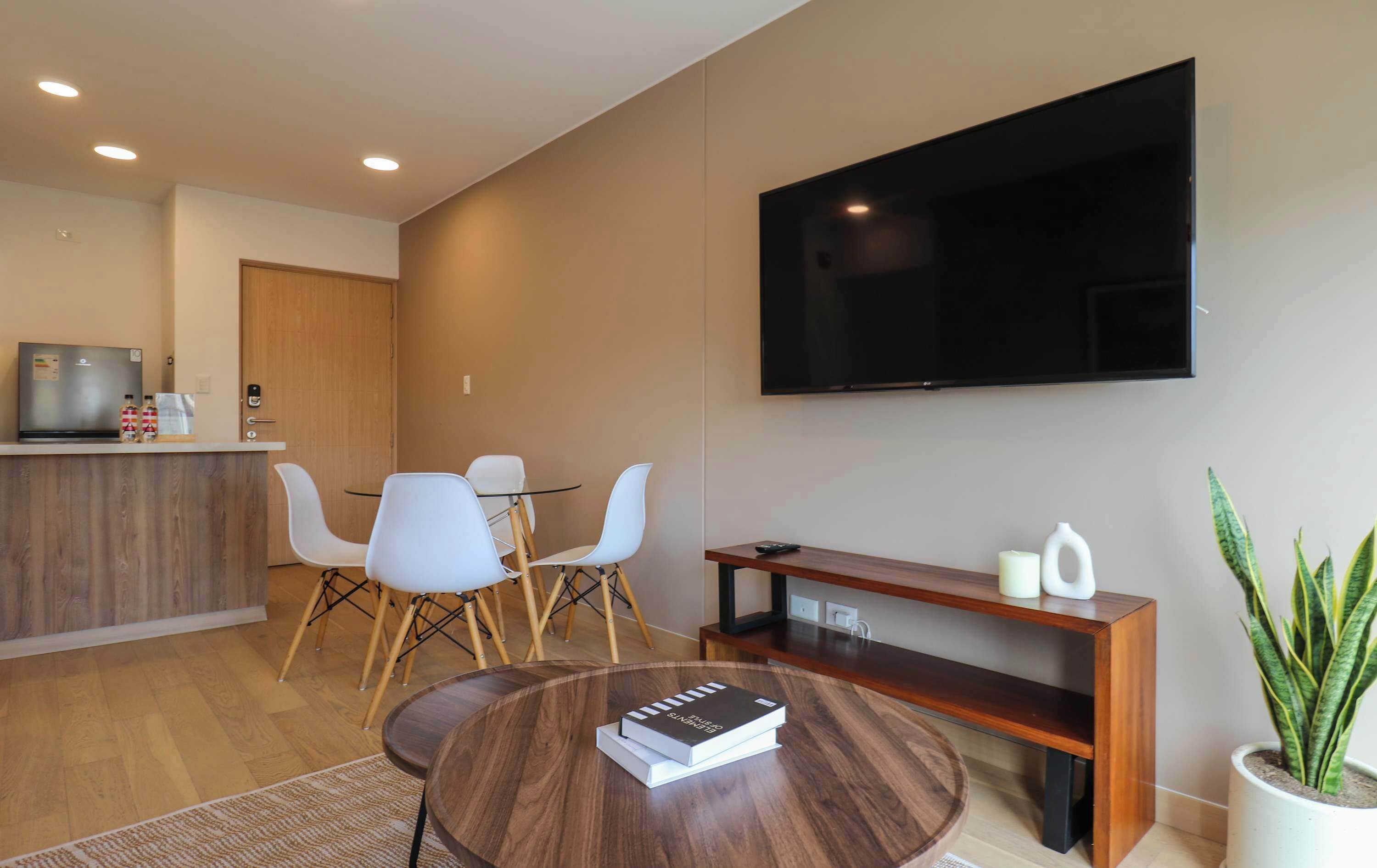 A modern living room with a flat-screen TV, a wooden coffee table with books, a dining area with chairs, and a potted plant.
