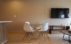 A modern living room with a round glass table, white chairs, a wooden bench, and a flat-screen TV mounted on the wall. - w* | Charming 2BR w/ Balcony & View in Miraflores (Miraflores)