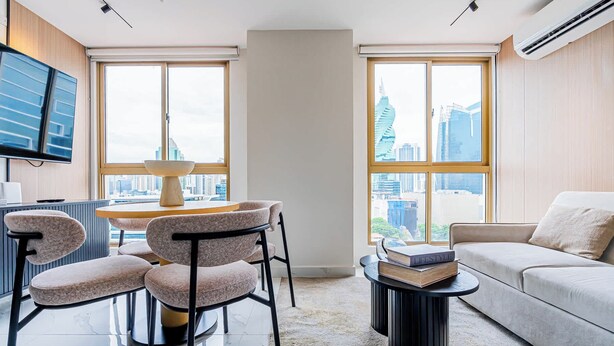 A modern living room with a sofa, a dining area with chairs, a TV, and a view of the cityscape through large windows. - w* | Upscale 1BR in Obarrio w/ AC (Panamá)