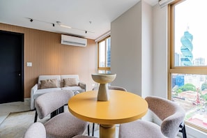 Ein modernes Wohnzimmer mit einem runden Tisch, einer Couch und Sesseln, einem großen Fenster mit Blick auf die Stadt und einer Klimaanlage an der Wand.