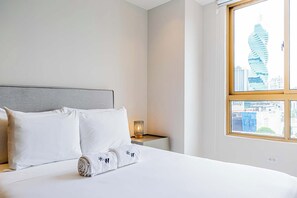 Ein Hotelzimmer mit Bett, Kissen, einem Nachttisch mit Lampe und einem Fenster mit Blick auf die Stadt.
