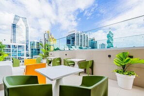 Et udendørs opholdsområde med borde og stole, plantekasser og udsigt til byens skyline.