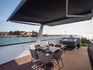 Eine Terrasse mit Tisch und Stühlen, einem Whirlpool und Blick auf Wasser und Gebäude.