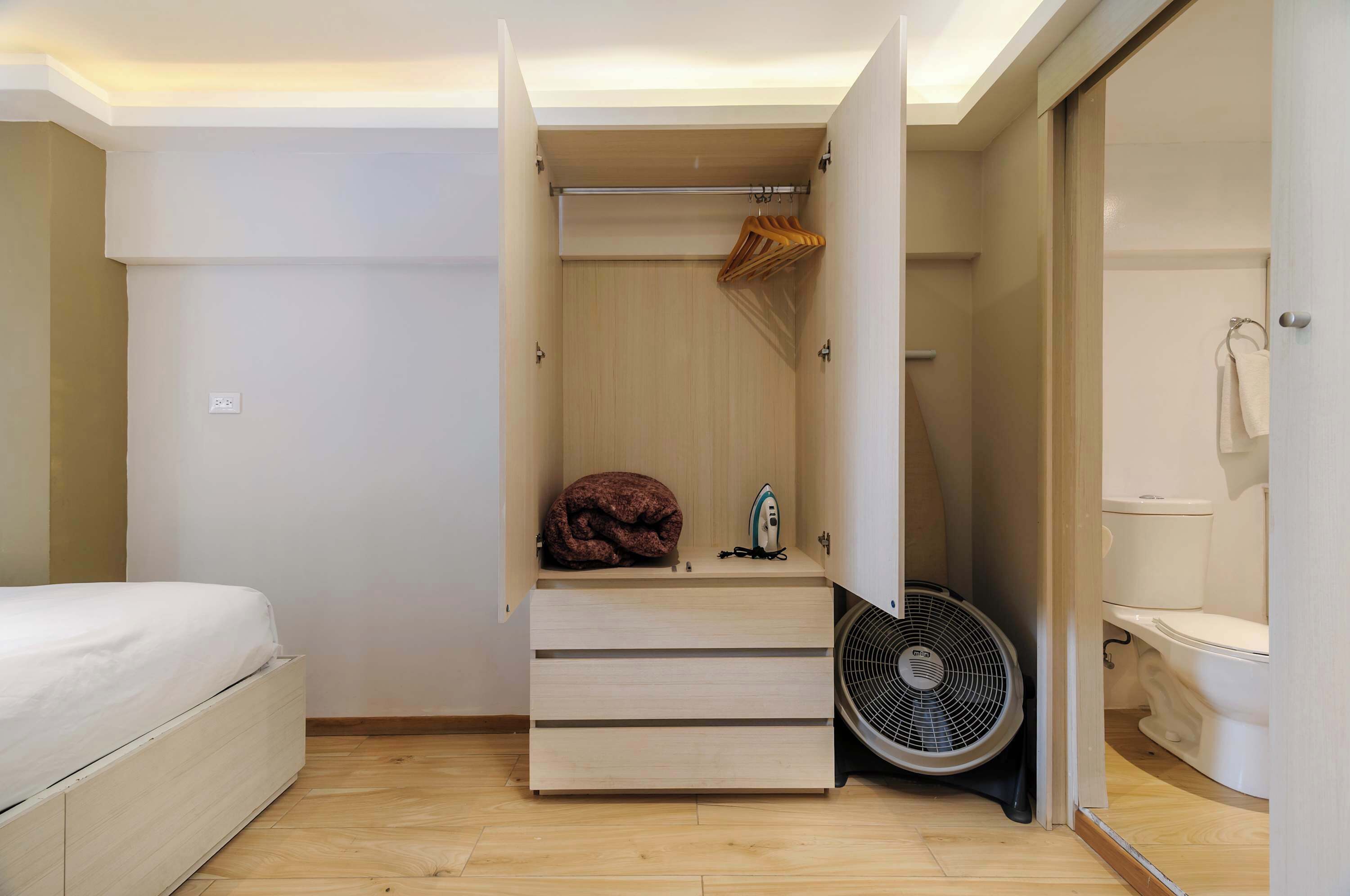 Une chambre avec un lit, une armoire avec une tringle pour suspendre les vêtements, un petit radiateur et un ventilateur.