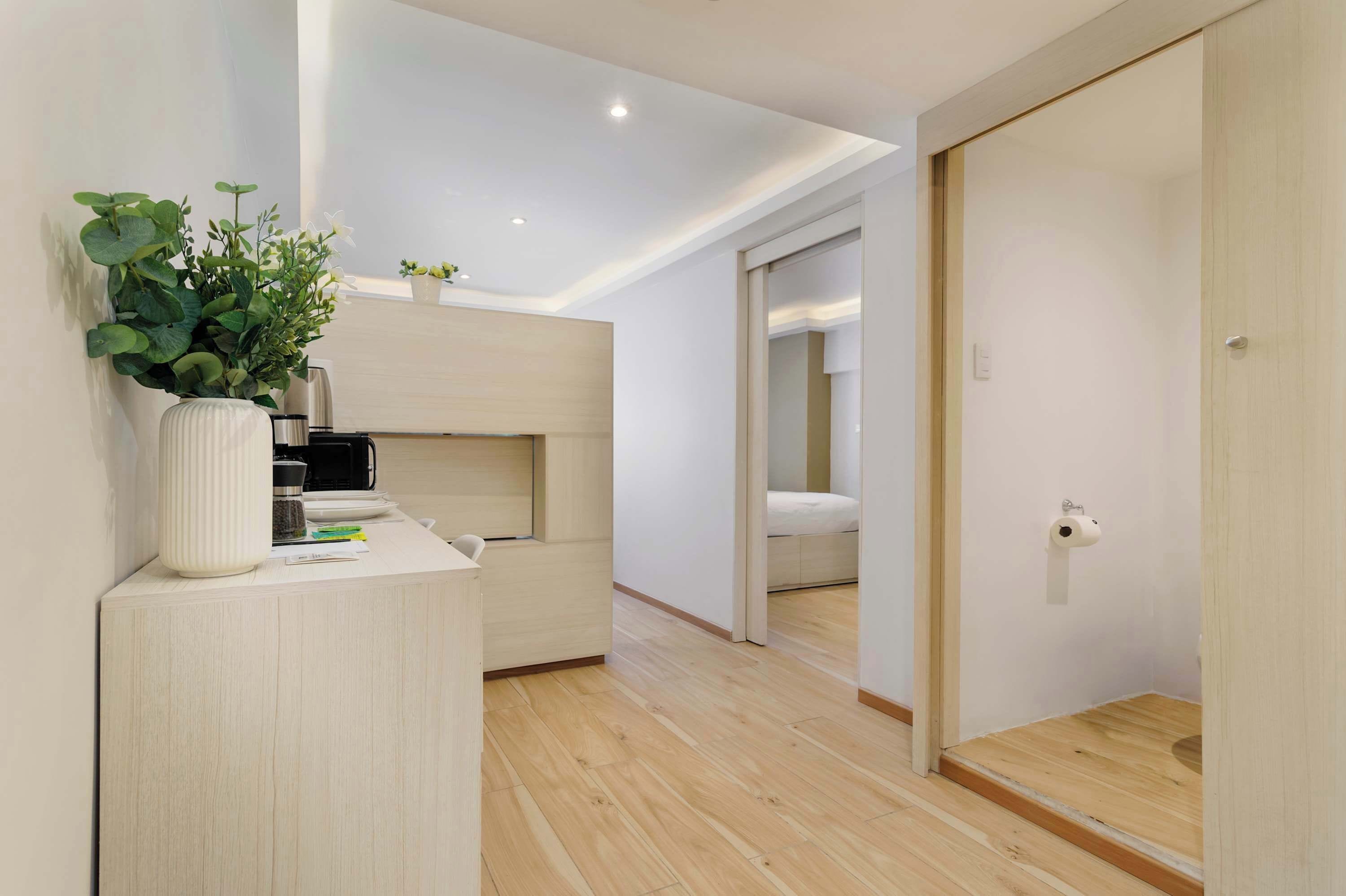 A modern, minimalist bedroom with a built-in bed, a bedside table with a plant, and a small desk with a kettle.