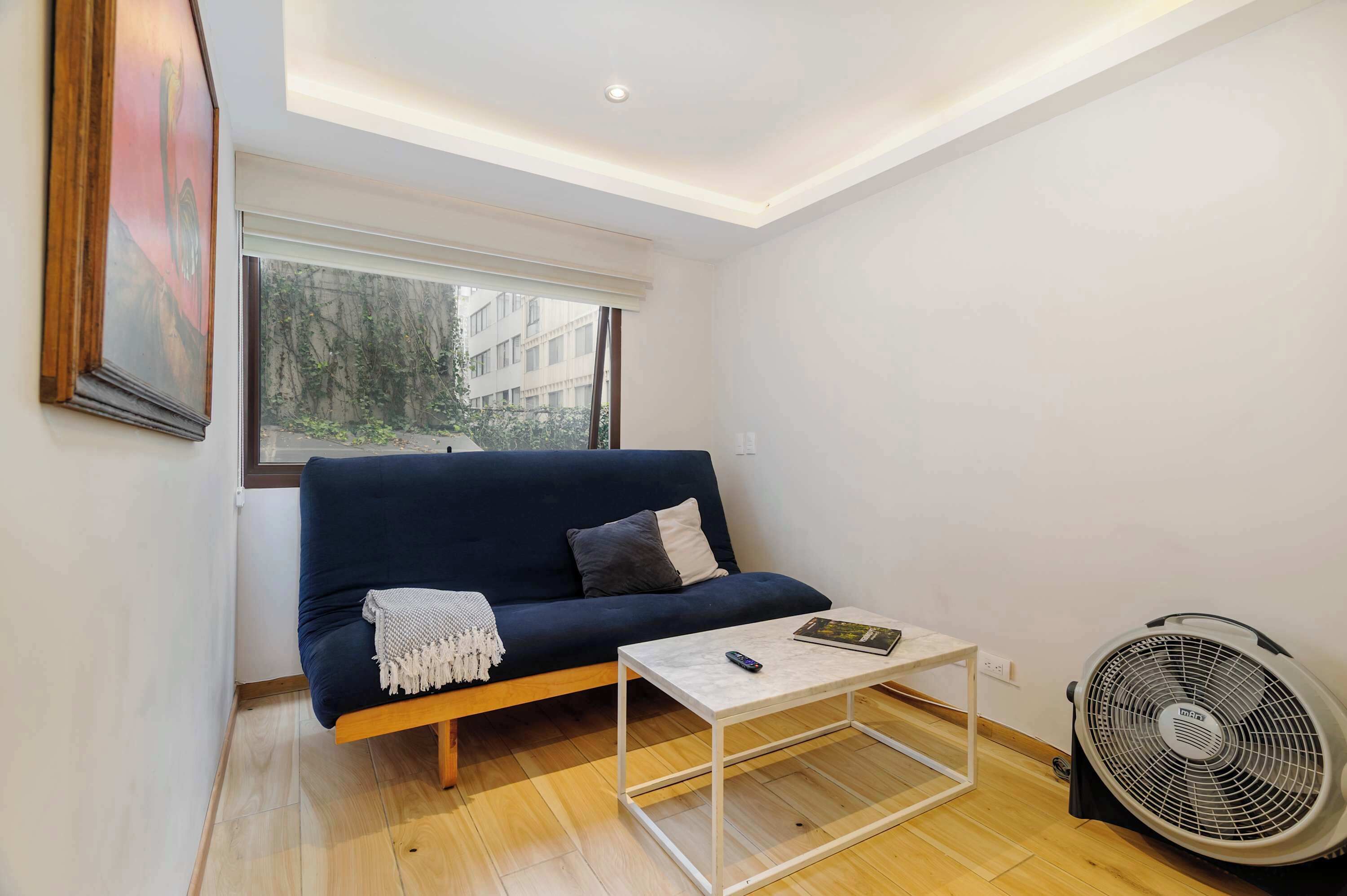 Un espacio habitable compacto con sofá, una mesita de centro, un ventilador y una ventana que ofrece vistas a edificios y zonas verdes.