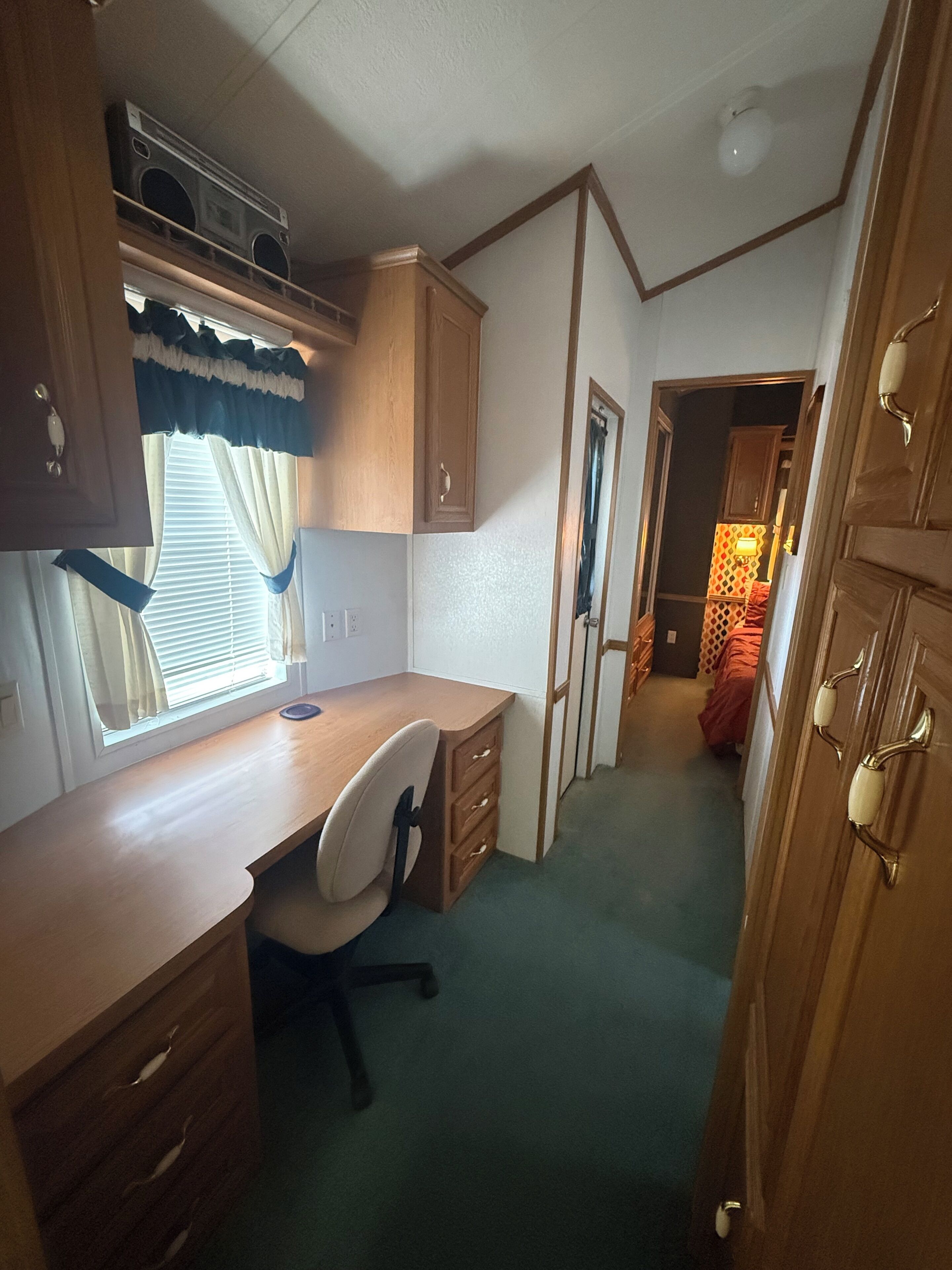 A compact room with a wooden desk, a chair, and a window with curtains. There is a door on the right and a wall-mounted light fixture.