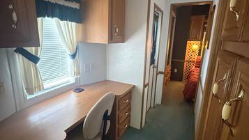 A compact room with a wooden desk, a chair, and a window with curtains. There is a door on the right and a wall-mounted light fixture.