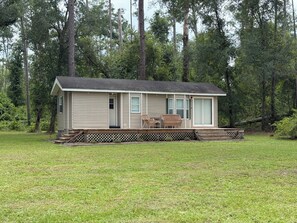 A single-story cabin with a porch, a small deck, and a lawn in a wooded area.