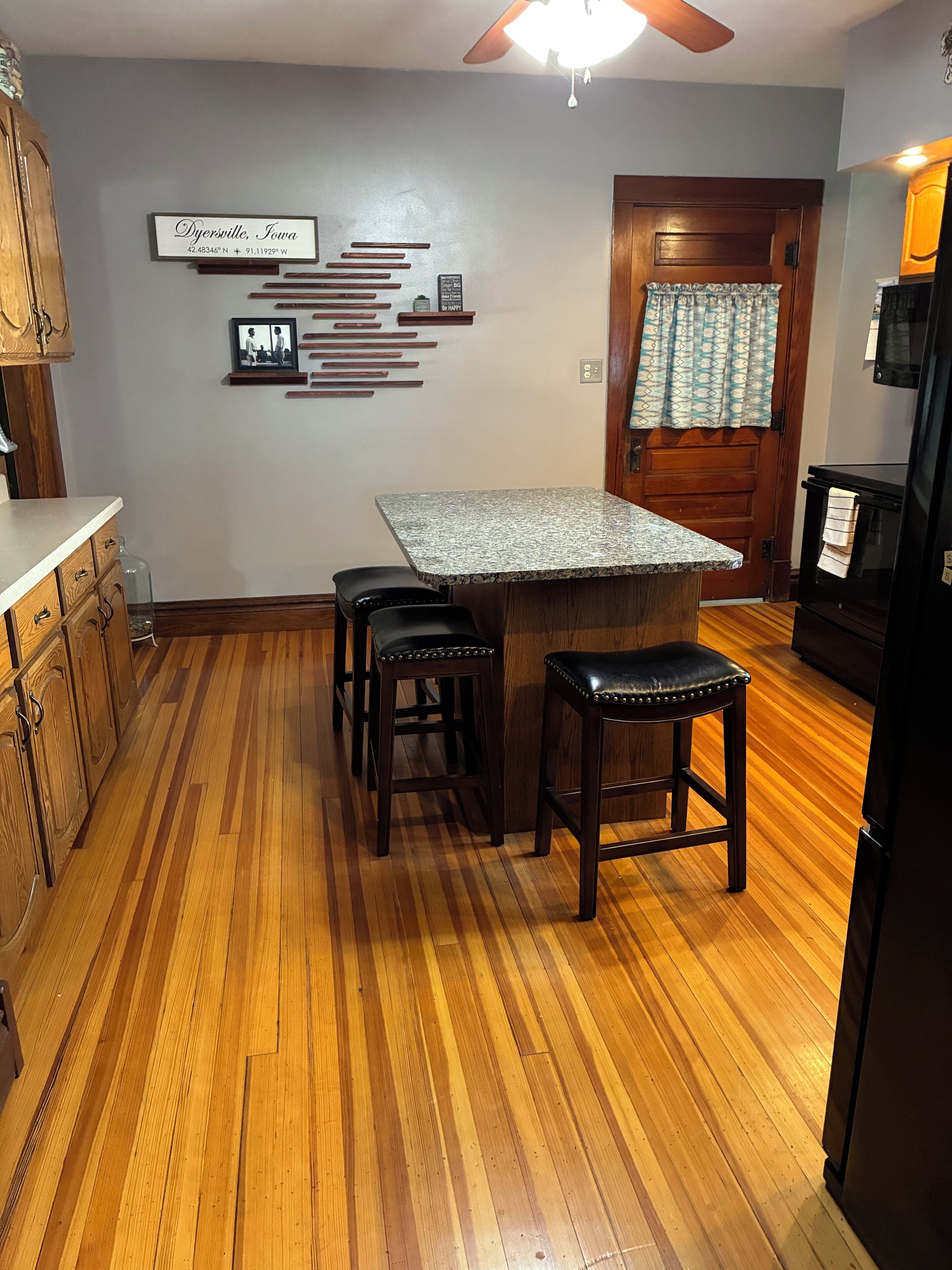 A kitchen with a wooden floor, a granite countertop island, two stools, and a refrigerator.