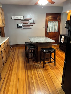 A kitchen with a wooden floor, a granite countertop island, two stools, and a refrigerator.