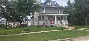 A two-story house with a front porch, a small front yard, and a flag commemorating an anniversary.