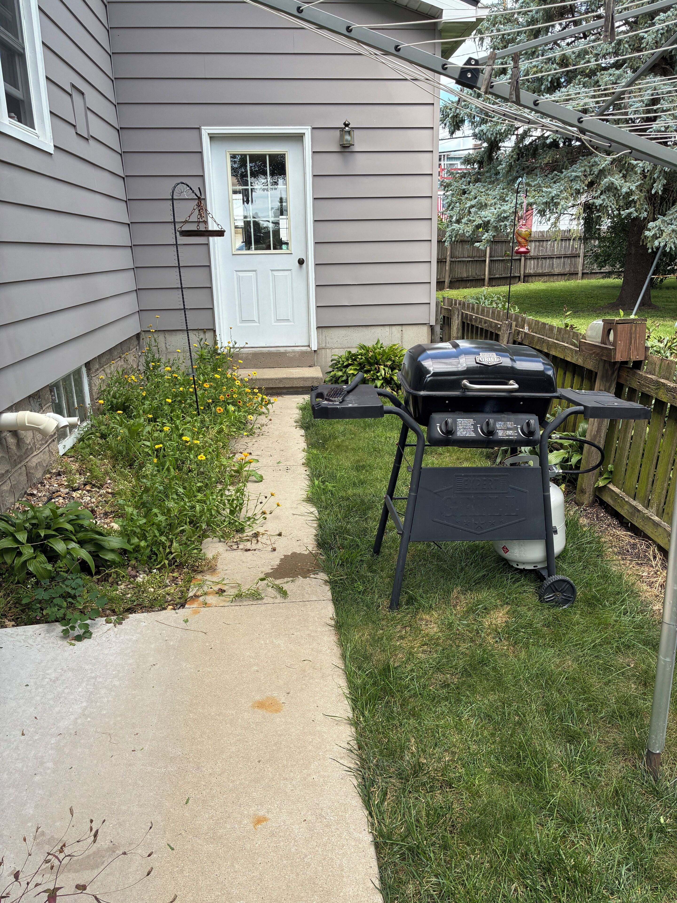 Un jardin avec un barbecue, un chemin et une maison avec une porte blanche.