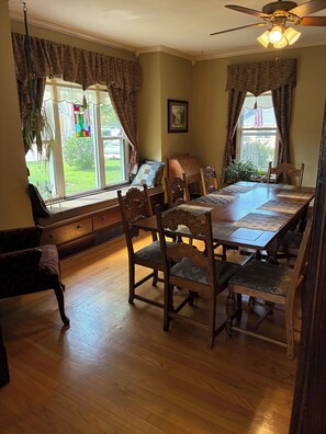 A dining room with a wooden table and chairs, a ceiling fan, and large windows with curtains.