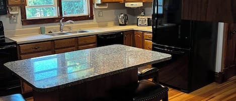 A kitchen with wooden cabinets, a black refrigerator, a granite countertop island with stools, and a ceiling fan.