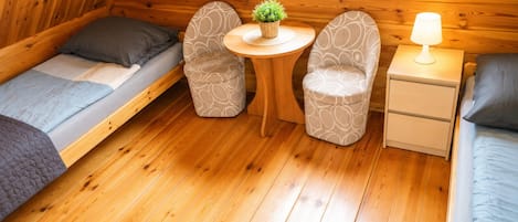 Habitación compacta con dos camas individuales, una mesita redonda con una planta, dos sillas estampadas, una mesita de noche con lámpara y paredes de madera.