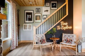 Ein gemütliches Interieur mit einer Treppe, zwei Korbstühle mit gemusterten Kissen, einem kleinen Holztisch und einem Neonzeichen mit der Aufschrift 'Time To Explore'.