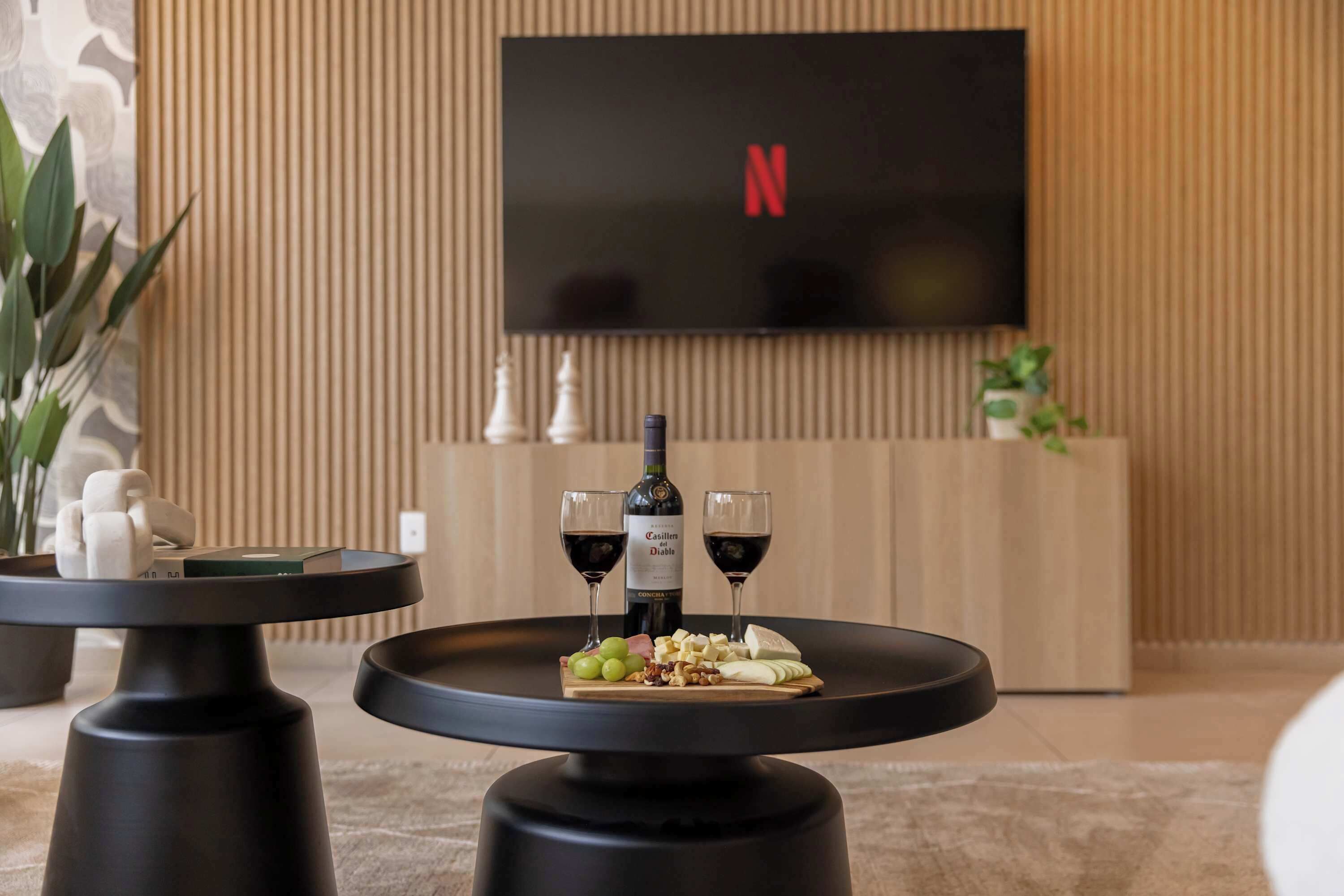 Una sala de estar moderna con un televisor de pantalla plana montado en la pared, una mesa redonda negra con una botella de vino y copas, y un aparador de madera con adornos.