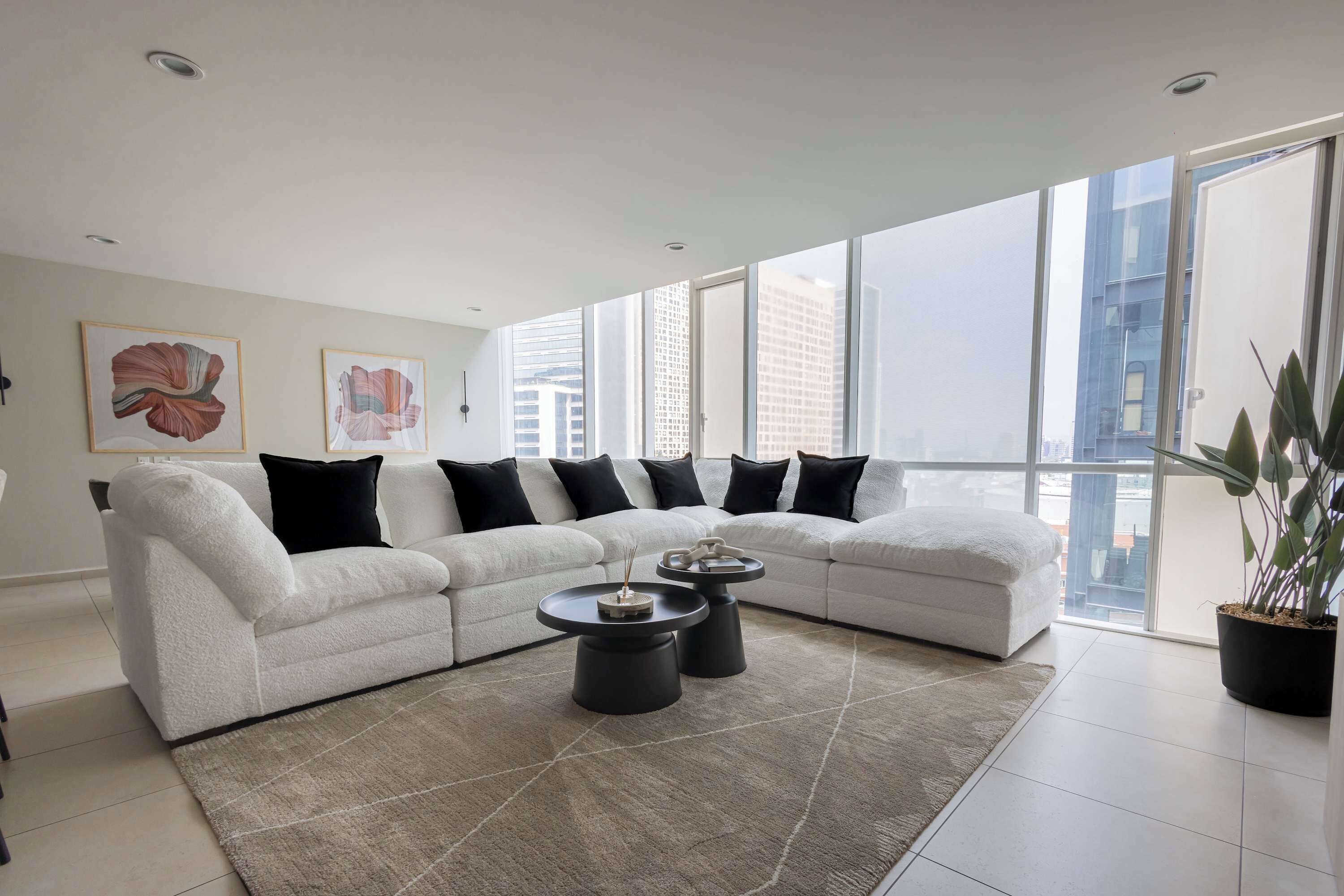 Ein modernes Wohnzimmer mit einem Ecksofa, zwei Beistelltischen und einem großen Fenster mit Blick auf die Stadt.