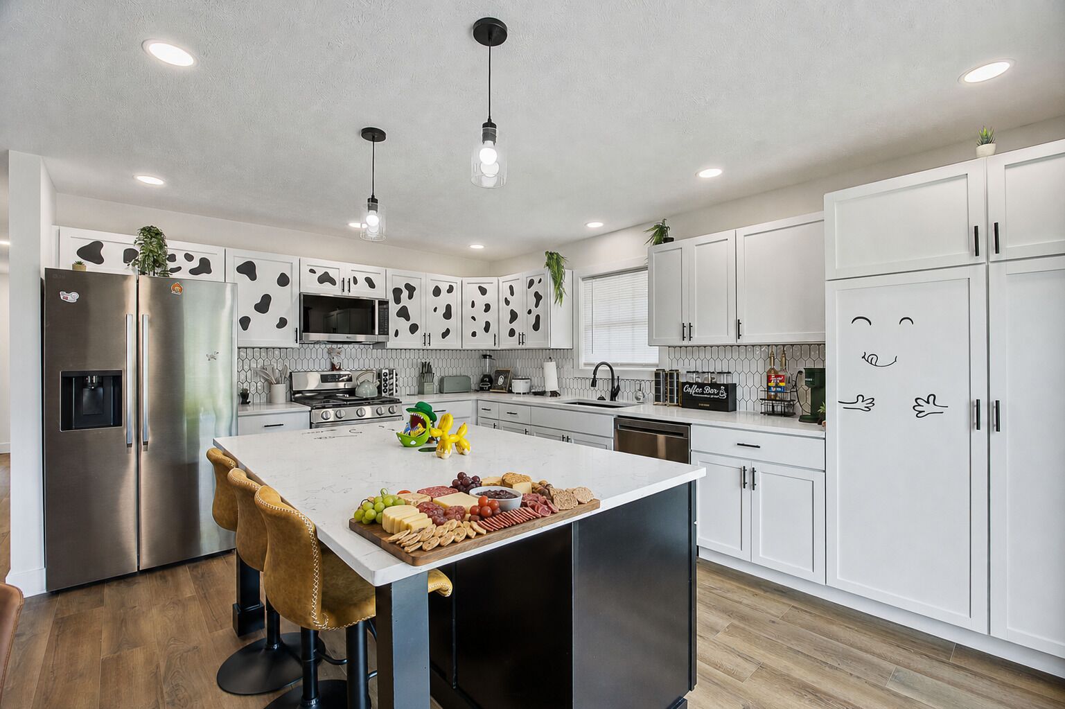 Cocina moderna con armarios blancos, electrodomésticos de acero inoxidable y una isla central con una tabla de quesos y frutas.