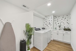 A laundry room with a washing machine, dryer, ironing board, and a potted plant.