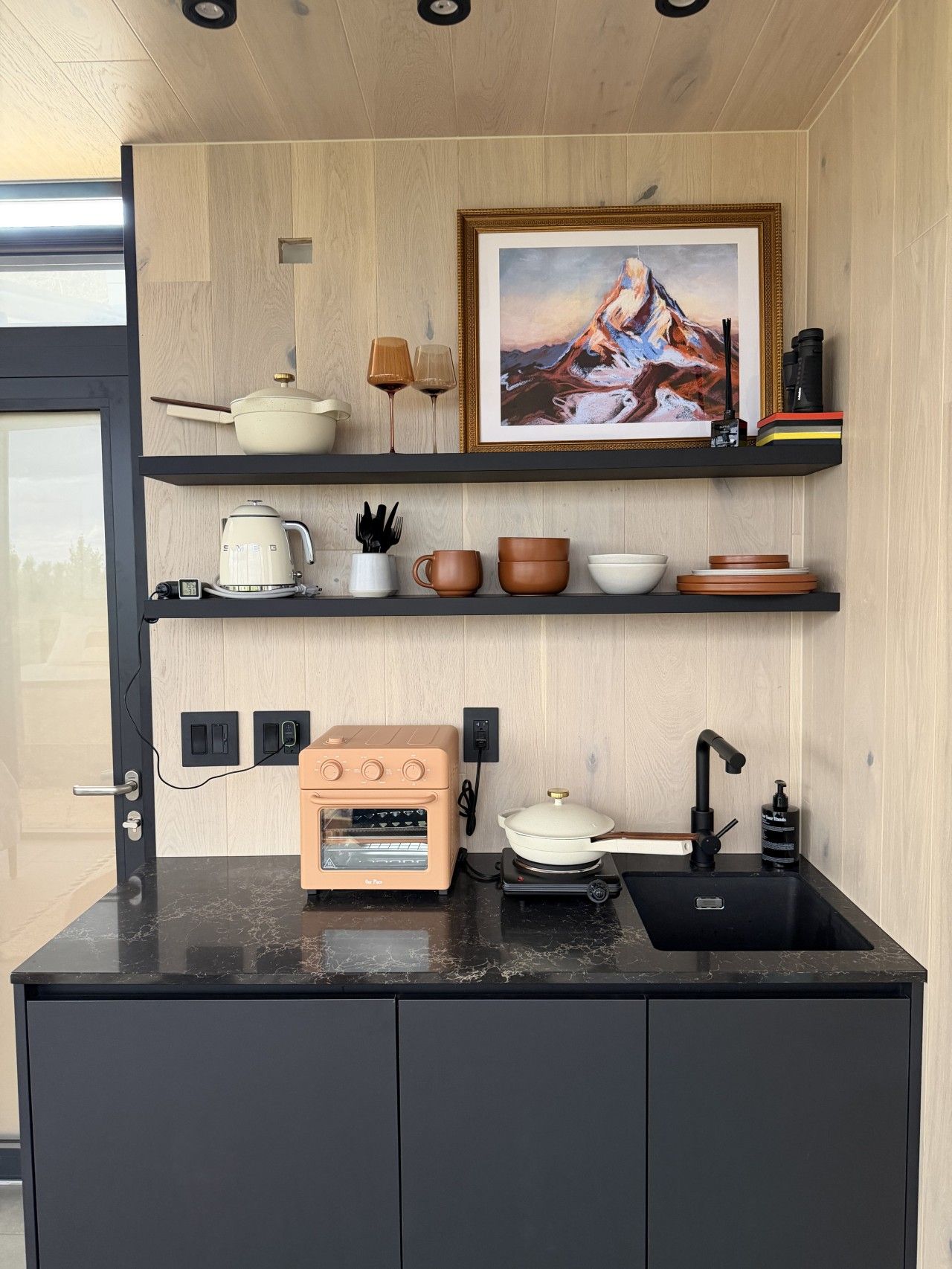 Cozinha moderna com forno embutido, pia e bancada. Há prateleiras sobre a bancada com diversos utensílios de cozinha e uma moldura com um quadro.