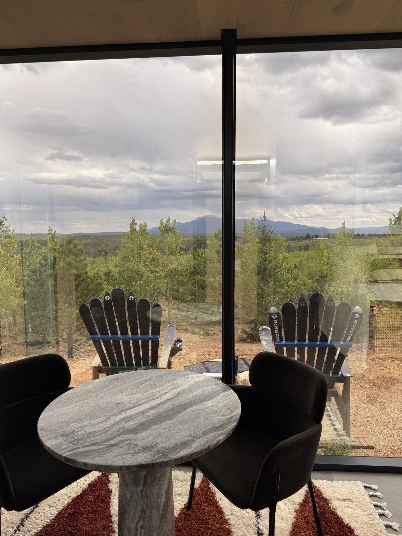 Quarto moderno com uma grande janela, duas cadeiras Adirondack, uma mesa redonda de mármore e vista para uma floresta e montanhas.