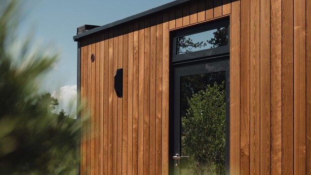 Un edificio moderno de madera con un amplio ventanal y puerta de vidrio, rodeado de follaje.