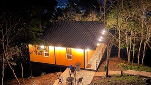 Een klein, laag huis met schuin dak, omringd door bomen en een gazon. Er is een klein terras met een tafel en stoelen.