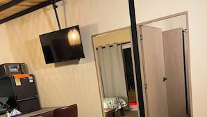Een compacte kamer met kitchenette, televisie aan de muur, bed met gordijn en een ronde tafel met afstandsbediening.