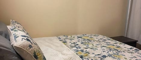 A bed with a floral quilt, a bedside lamp, and a grey sofa in a room with plain walls.