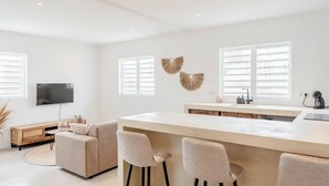 A modern kitchen with a central island, bar stools, a flat-screen TV, and decorative wall art. - Kas Leon (Willemstad)