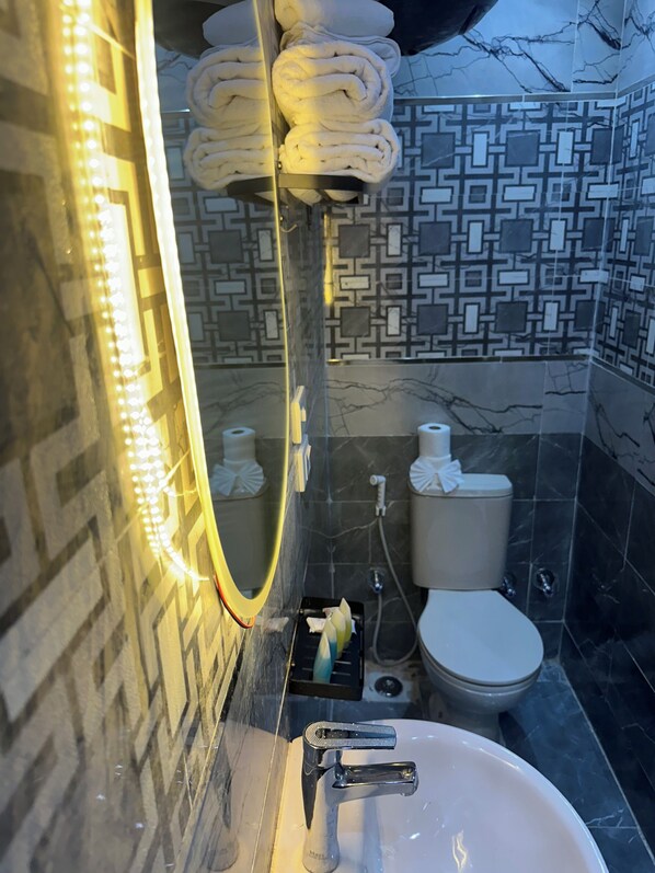 Un baño con pared de azulejos de mármol, un lavamanos blanco con un solo grifo, un inodoro y un perchero con toallas dobladas.