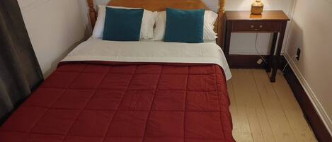 Een bed met een houten hoofdbord, witte en teal kussens en een rode sprei. Er staat een nachtkastje met een lamp en een lade.