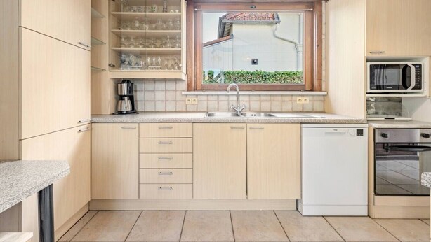 A kitchen with light wood cabinets, a built-in microwave, a dishwasher, and a window with a view of greenery. - Le Molinel - vakantiewoning in hartje Koksijde (Koksijde)